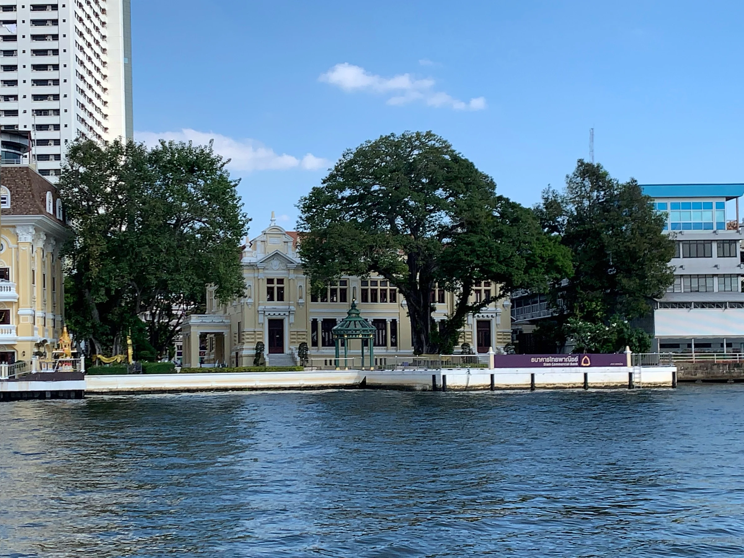 The cream-coloured Siam Commercial Bank Talat Noi branch building behind large trees on the bank of the Chao Phraya River in Bangkok under a blue sky