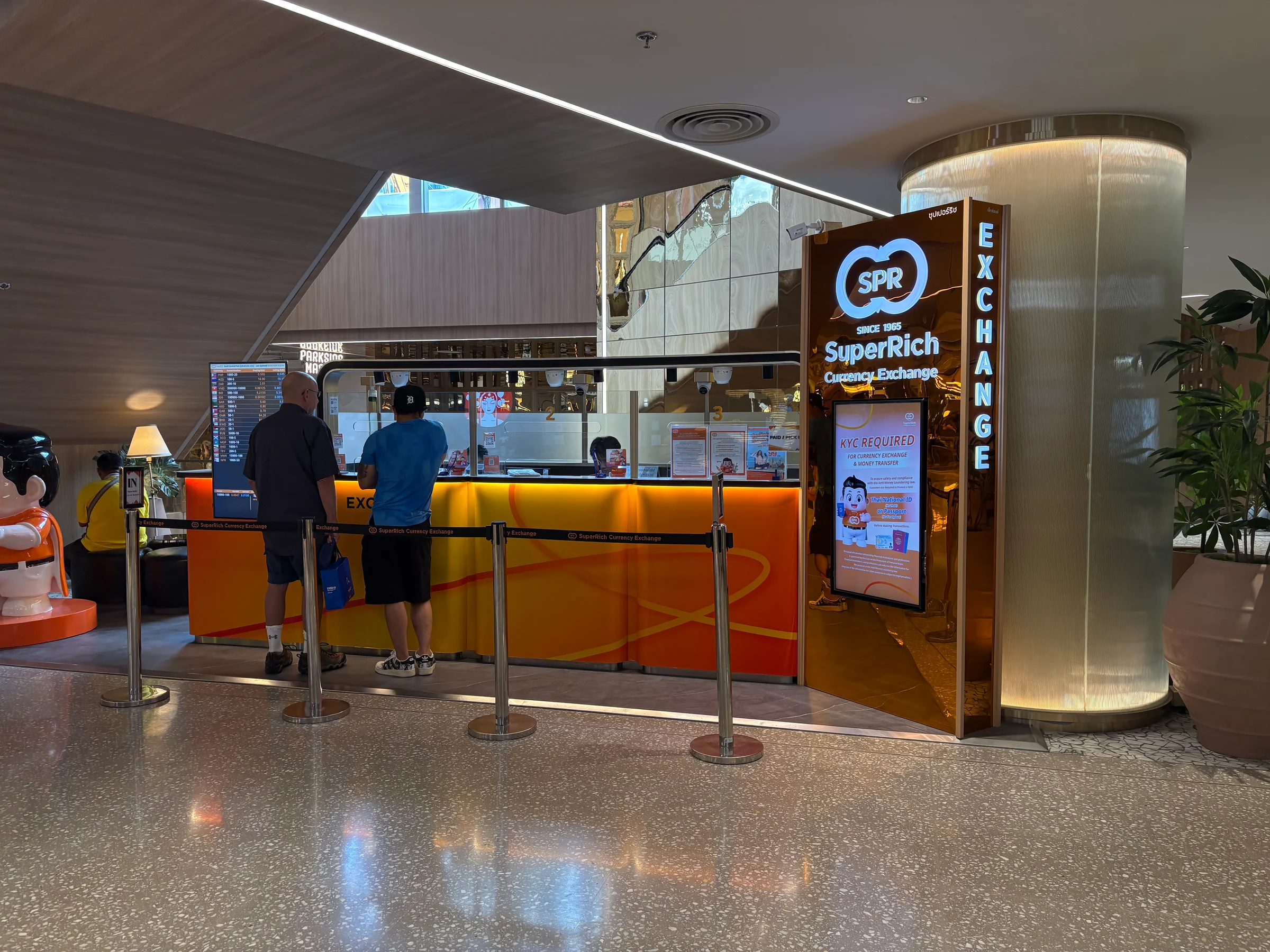 Customers at an orange-lit currency exchange counter in a Bangkok shopping centre, beside a gold sign reading Exchange