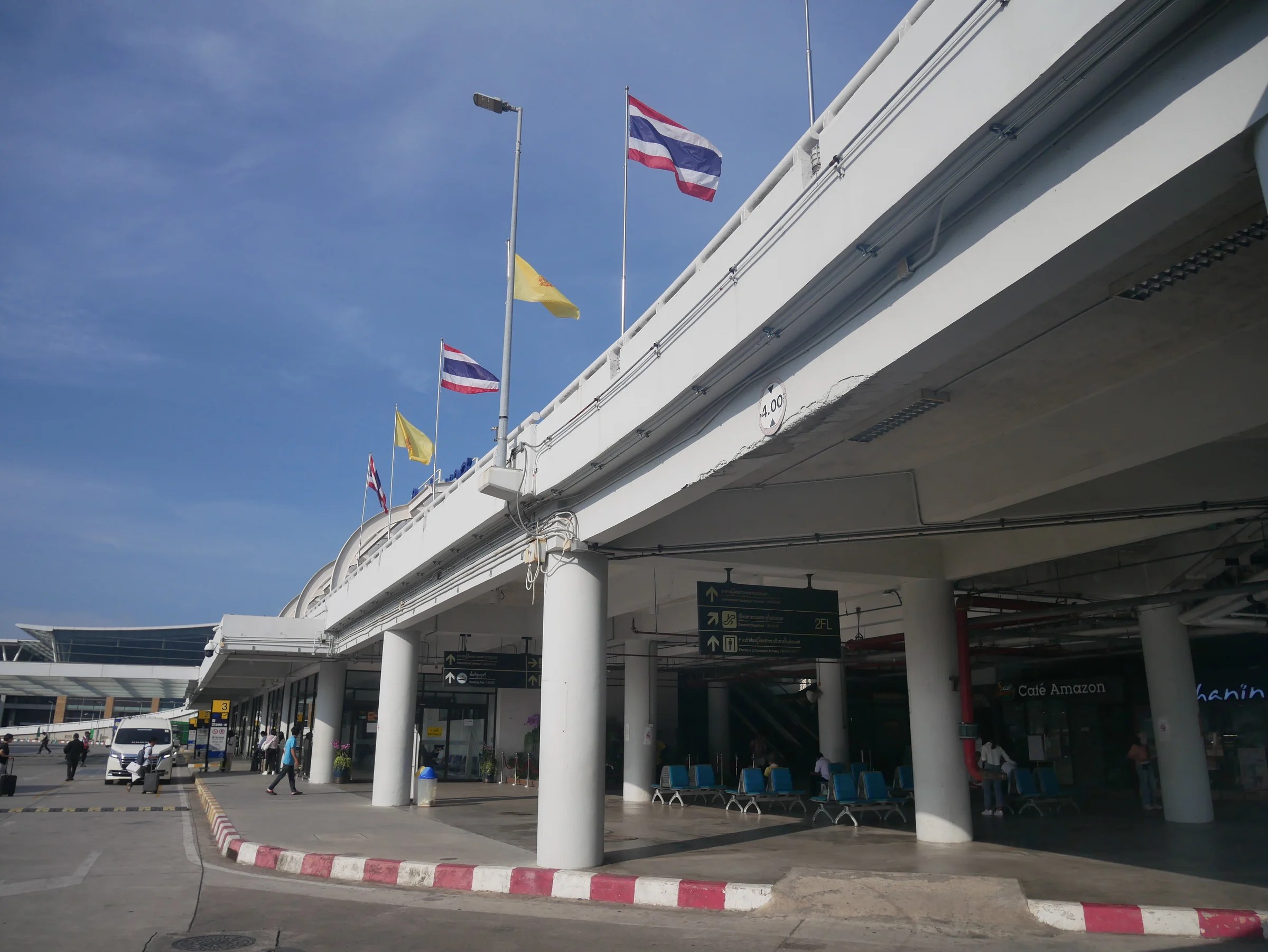 Departures frontage of Phuket International Airport under a blue sky, with Thai national and royal flags above the elevated drop-off road