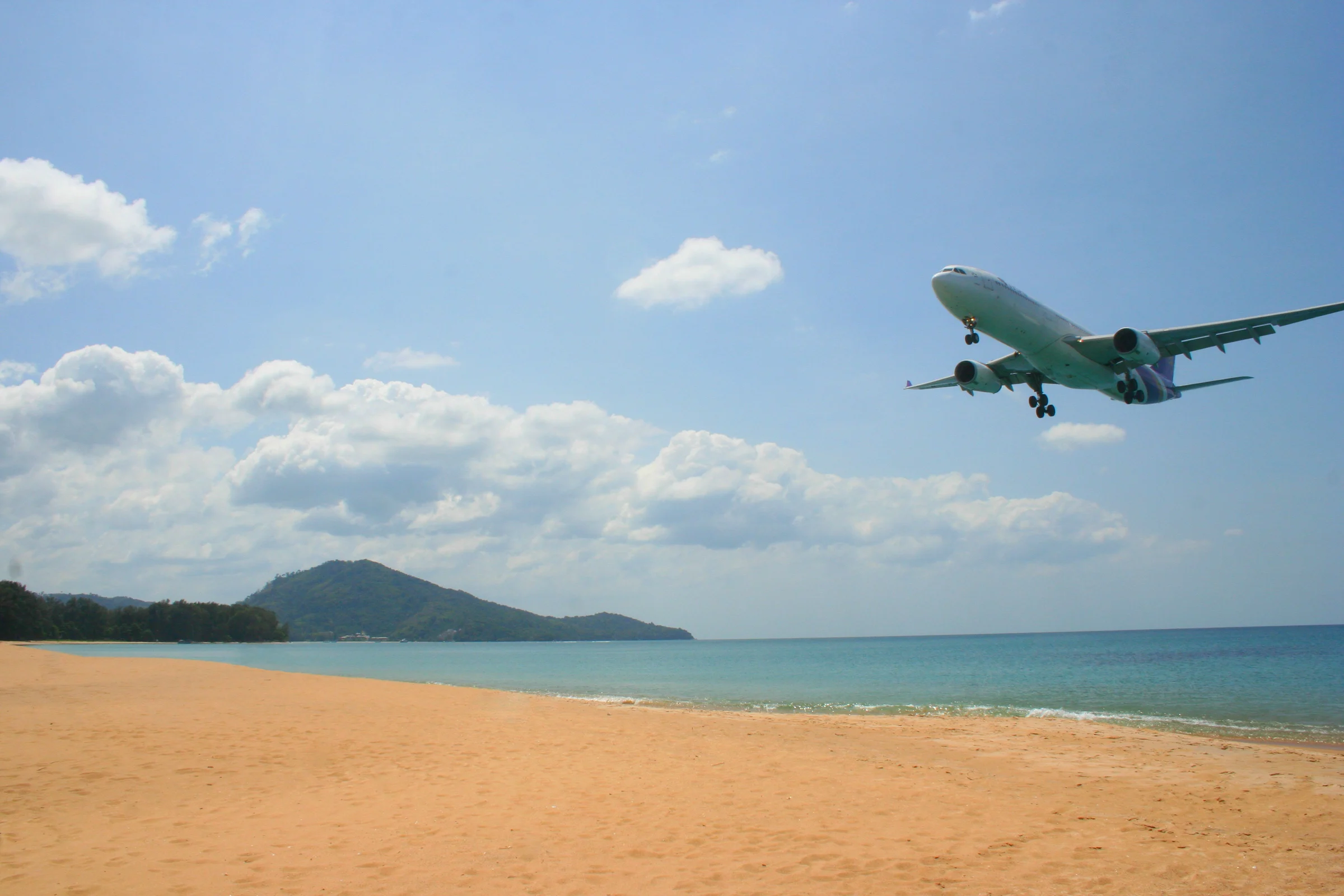 A twin-engine airliner on final approach low over the sand of Mai Khao Beach in Phuket, with a forested headland and the Andaman Sea behind