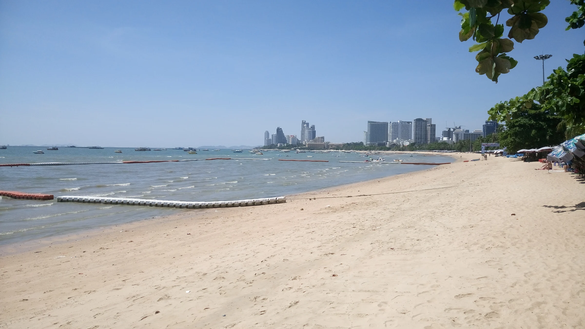 A wide sandy stretch of Pattaya Beach under a clear blue sky, with calm water, swimming area buoys and the Pattaya hotel skyline across the bay