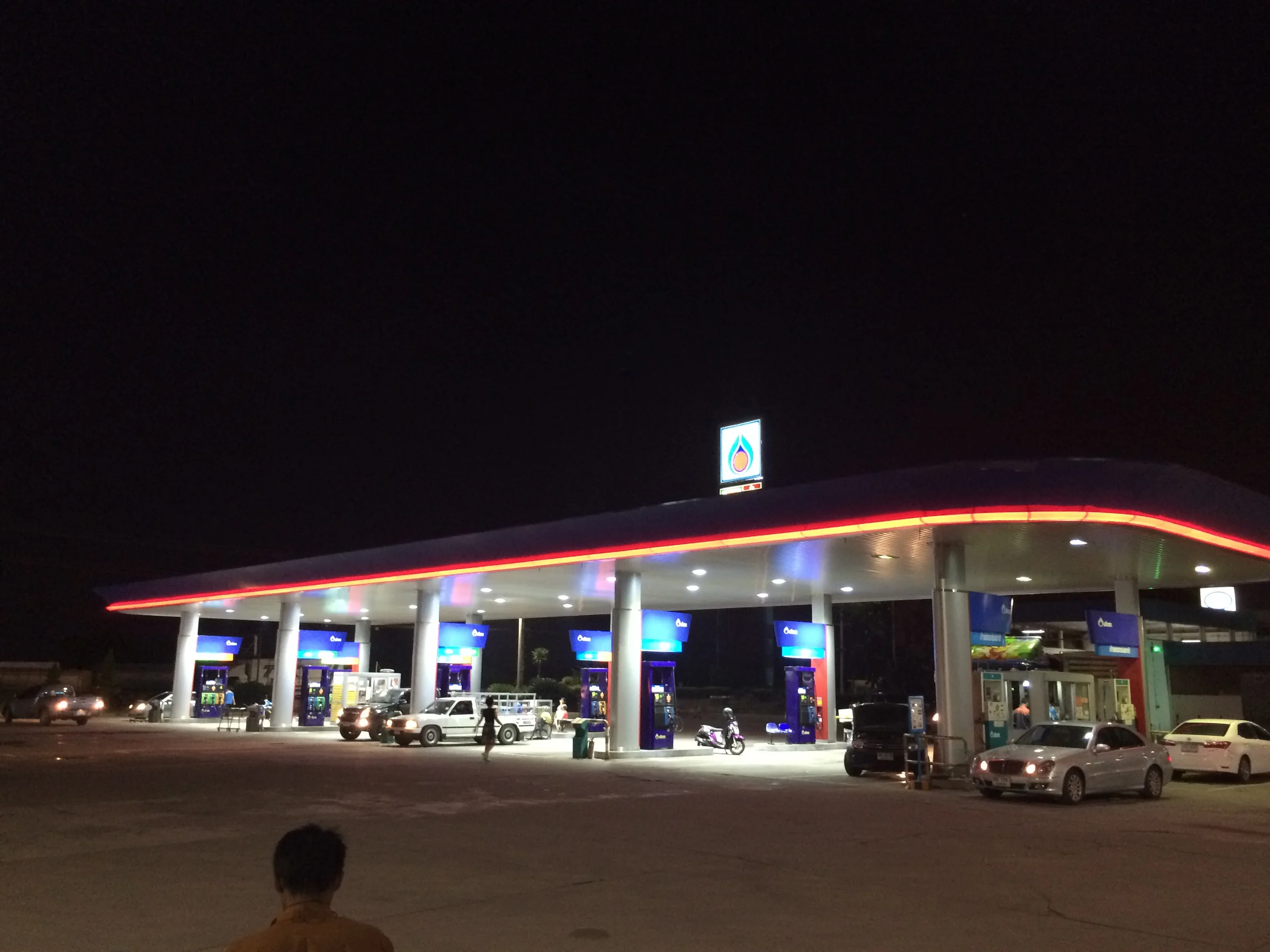 A PTT filling station lit up at night in Samut Songkhram province, with a red and orange lit canopy, blue fuel pumps and cars on the forecourt