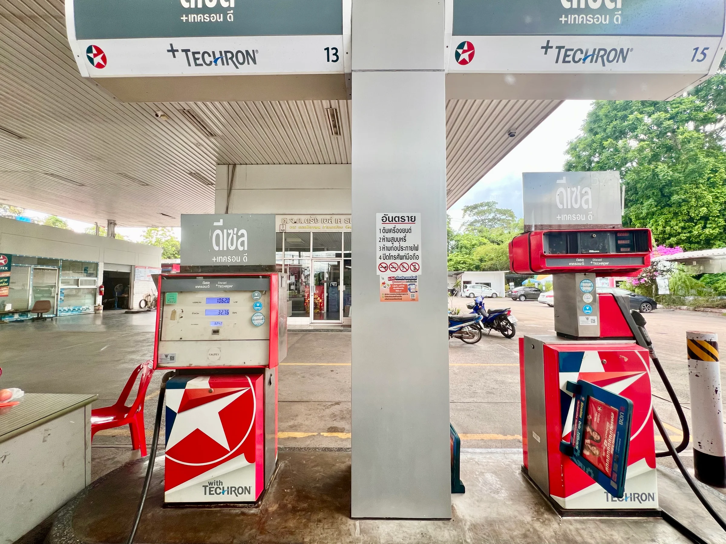 Red and white diesel pumps under the canopy of a petrol station forecourt in Trang, southern Thailand, with green trees behind