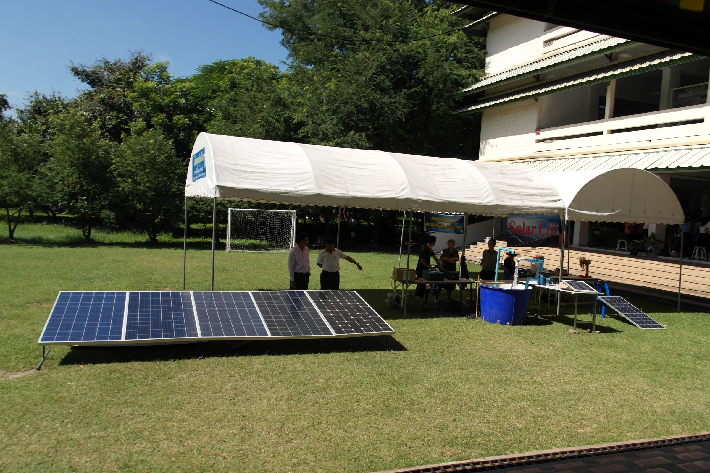 A row of blue solar panels on a green lawn in Thailand, with people at a demonstration tent and a white school building behind