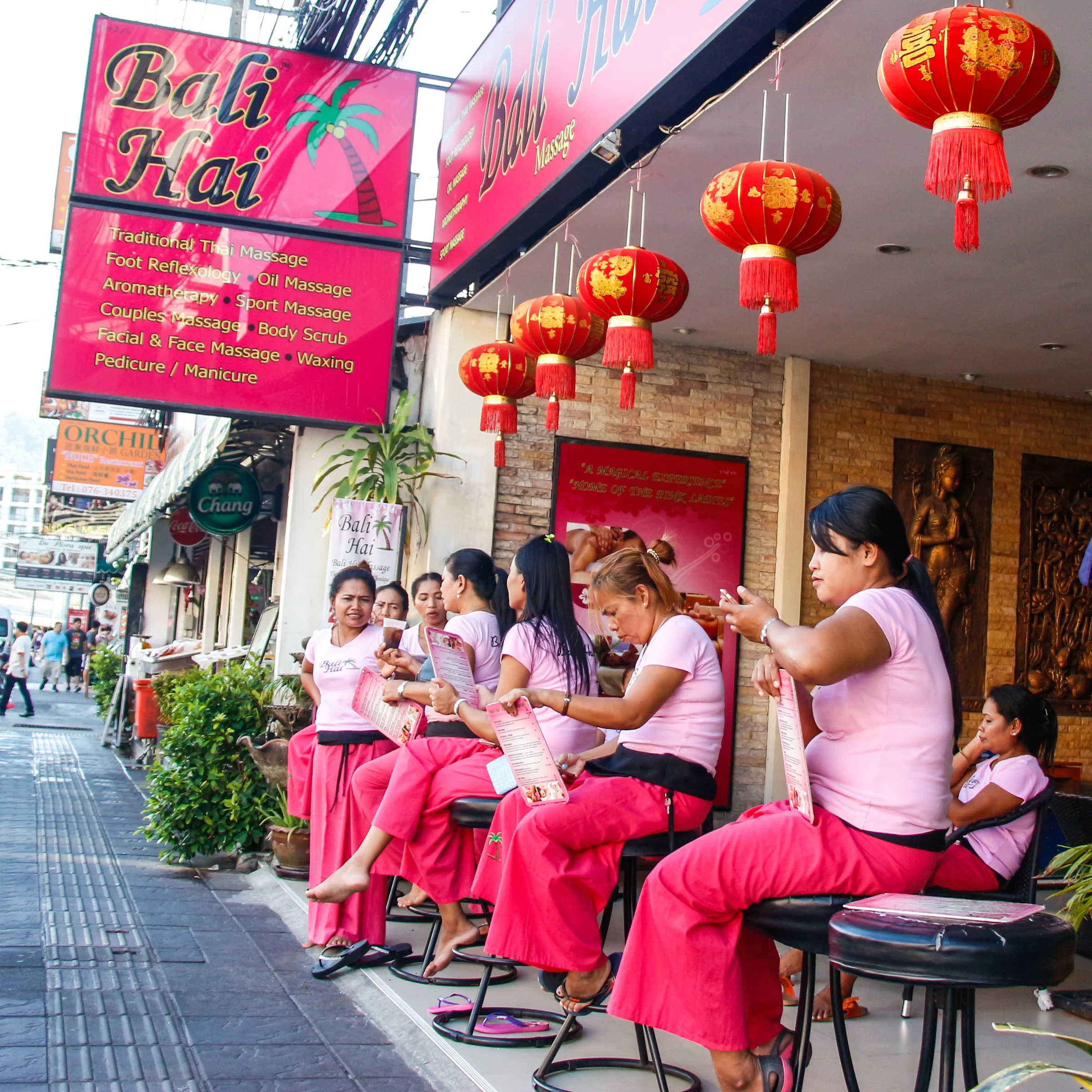 Massage therapists in pink uniforms seated outside a massage shop decorated with red lanterns on a street in Phuket
