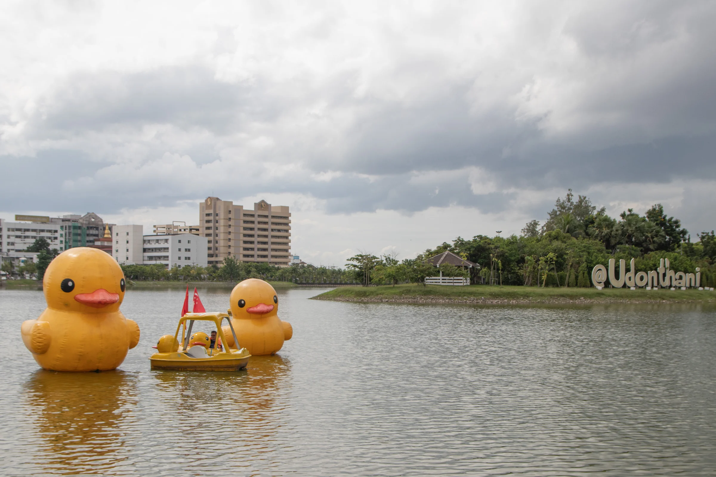 Giant yellow inflatable ducks and a duck pedal boat on Nong Prajak lake in Udon Thani, with green trees and an Udonthani sign on the far bank